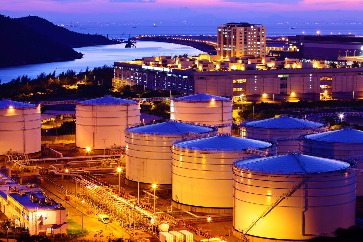 Aerial view of a coastal industrial facility at dusk, featuring large illuminated cylindrical storage tanks, refinery buildings, pipelines, a bridge over water, and a dark shoreline in the background.