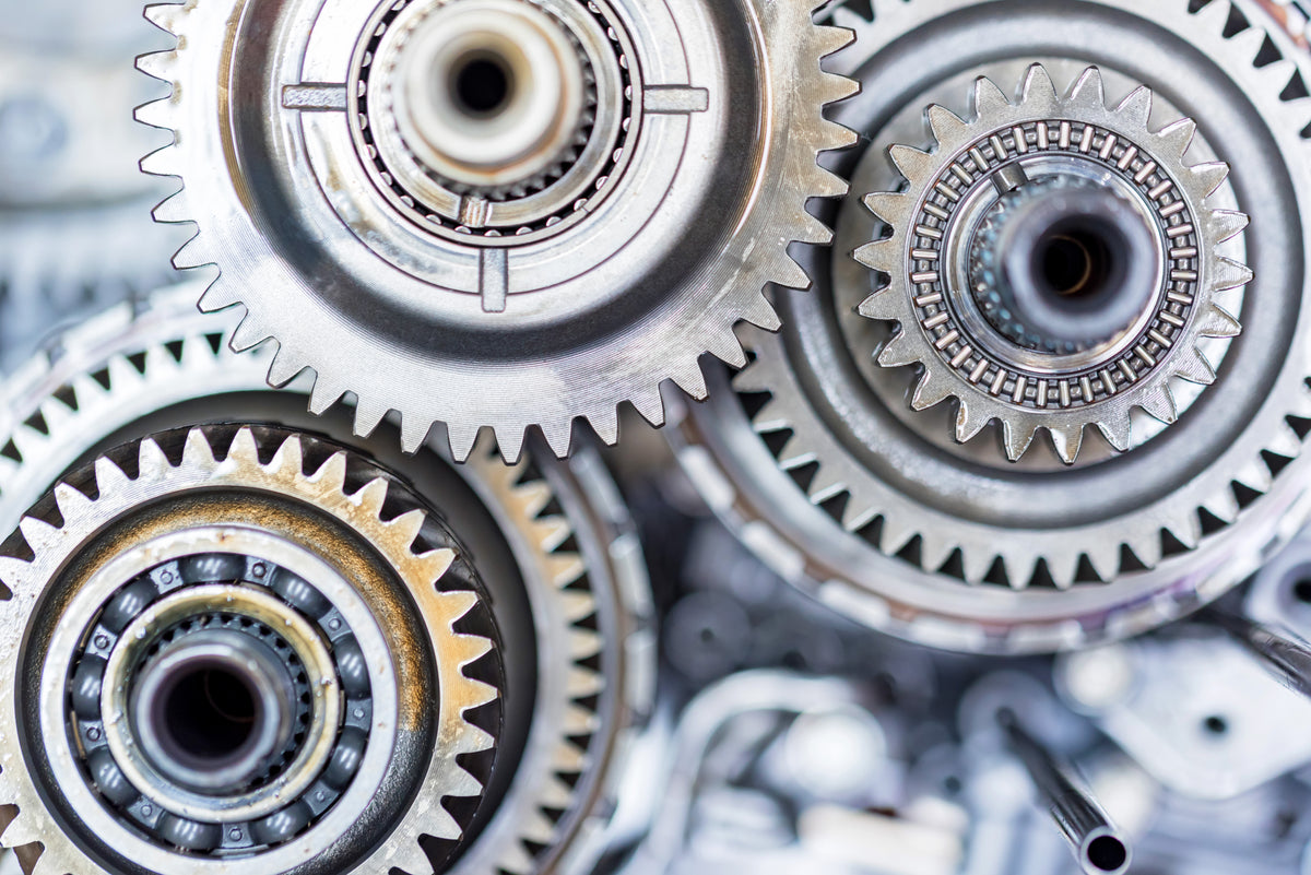 Close-up of metallic gears and sprockets with visible teeth, bearings, and industrial metal surfaces in silver and gray tones.